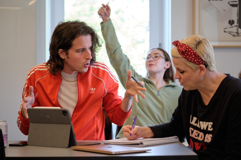 Students discuss a topic at their seats in Italian class. Another student raises her hand behind them. 