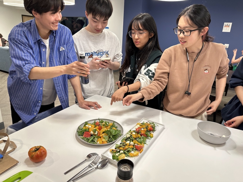 Students make a salad together, standing over the ingredients at a white table.