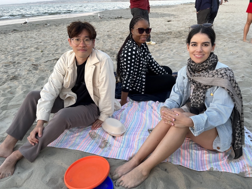 Students sit on a towel on the beach together, smiling at the camera. 