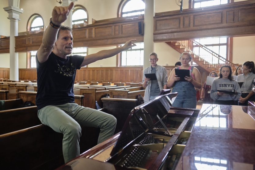 A man conducts a choir, sitting atop a pew. 