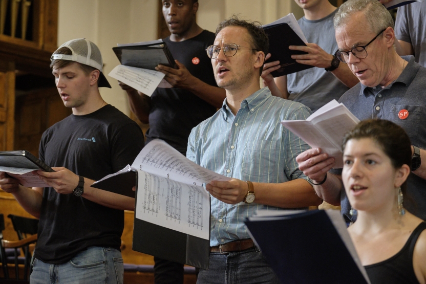 Adults sing in a choir, wearing pins saying "SP" on their chests. 