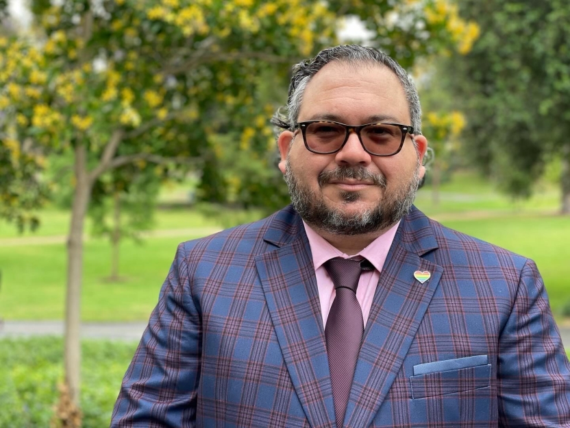 A man stands in front of green trees in a suit and tie. 