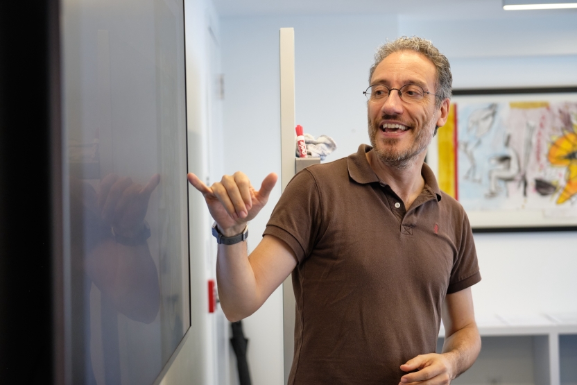 A teacher smiles as he gestures at a blackboard.