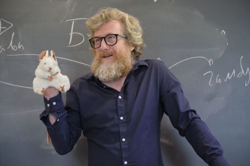 A man with a mouse puppet smiles at a blackboard with Russian writing on it. 