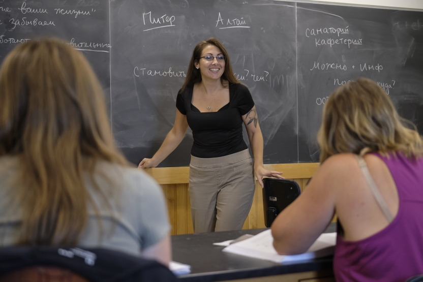 A teacher smiles at her students in front of a blackboard with Russian writing on it. 
