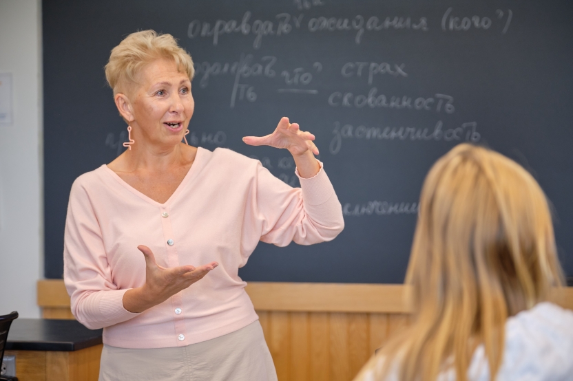 A teacher gestures at a blackboard, smiling at her students. 