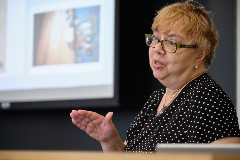 A woman in a black and white polka dot shirt speaks at a projector screen.