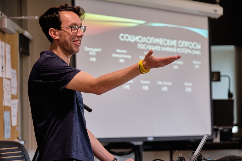 A man gestures, smiling at his students, standing in front of a projector screen.