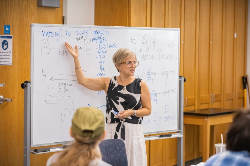 A woman in a black and white shirt and white skirt teaches at a white board.