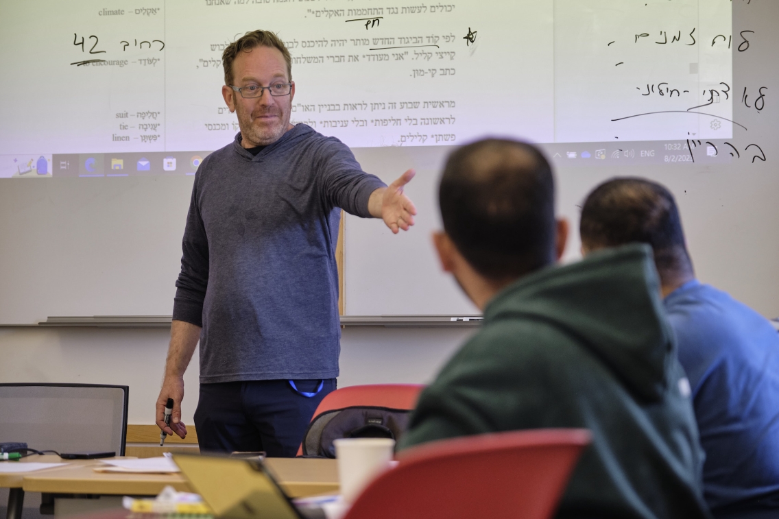 A teacher reaches his hands out to his students, awaiting an answer. He stands in front of a screen with Hebrew writing on it. 
