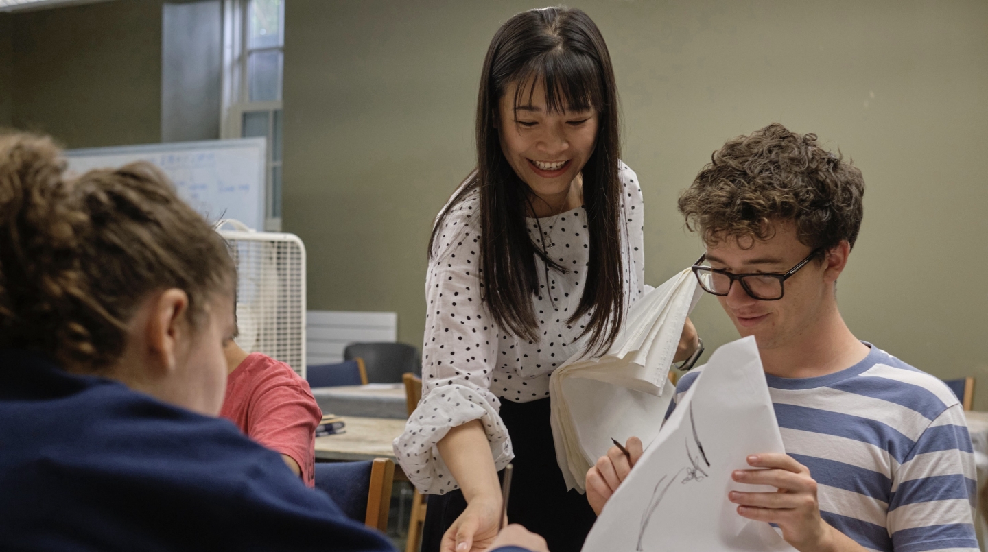 A teacher helps her student, standing behind him at his desk.