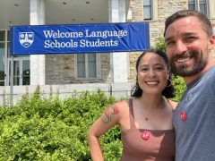 Two people smile together with red pins that say SP on their chests in front of a Welcome Language Schools Students Sign.