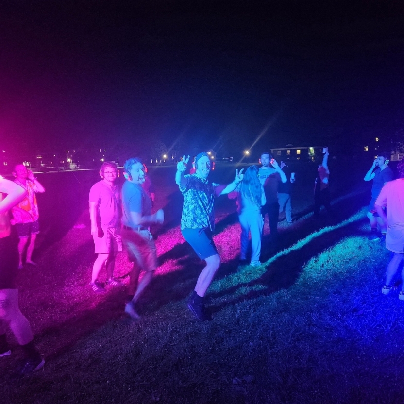 Students dance in the dark surrounded by colorful lights with headphones on. 