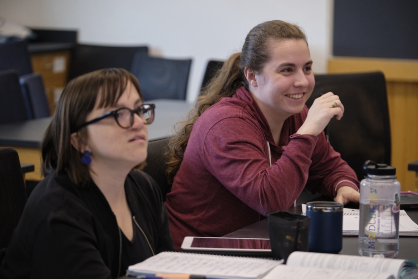 Two students sit together at their desk smiling as they listen to their professor.