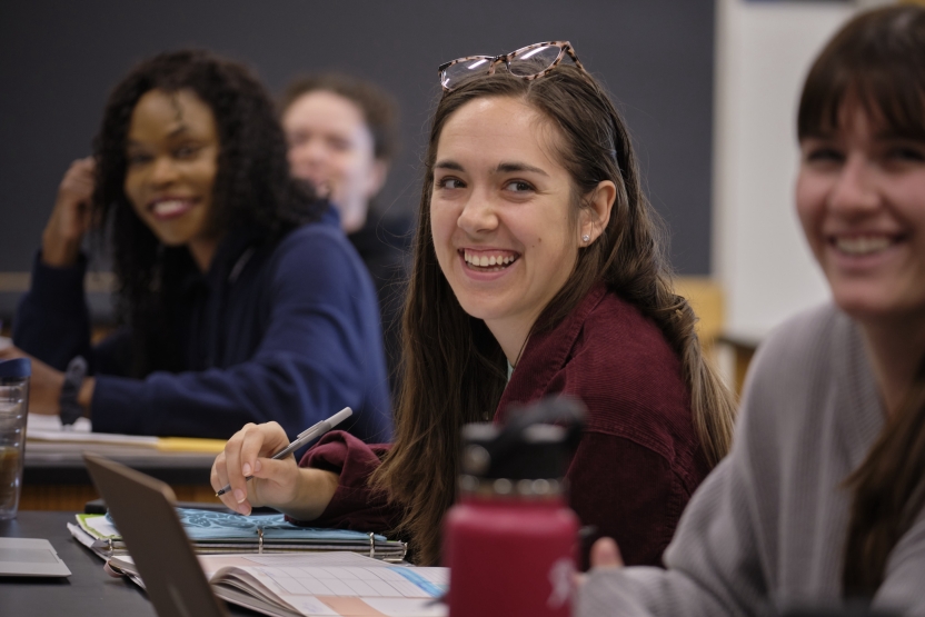 Three women smile as they look up at their teacher in class.