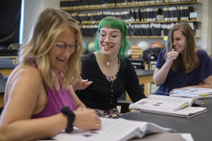 Three students laugh as they discuss a passage of a Russian book together at a table.