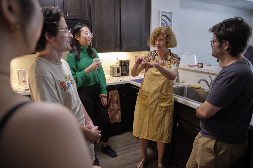 A woman acts out layering a cake to a group of people in a kitchen. 