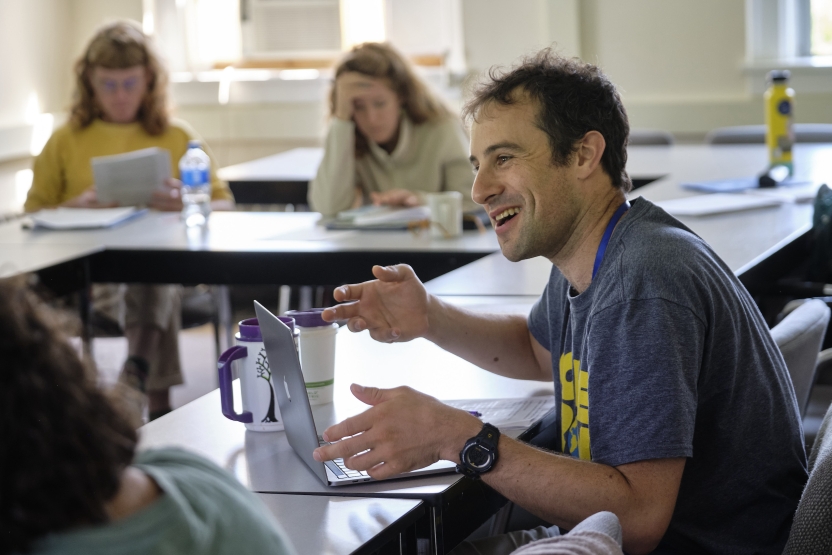 A student sits at a table in class, speaking and smiling.