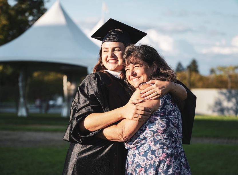 A mother hugs her daughter as she graduates with her Master's degree in front of a white tent.