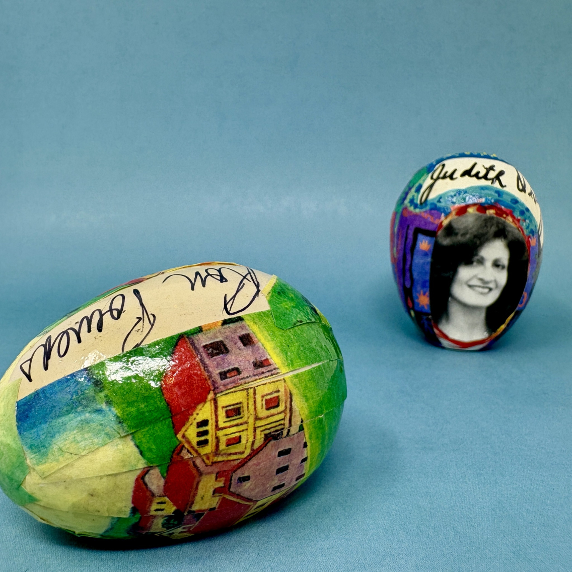 two wooden eggs decorated with collage and red, orange, purple, yellow, blue, and green paintings and collage, signed by Ron Powers and Judith Ortiz Cofer against a blue background