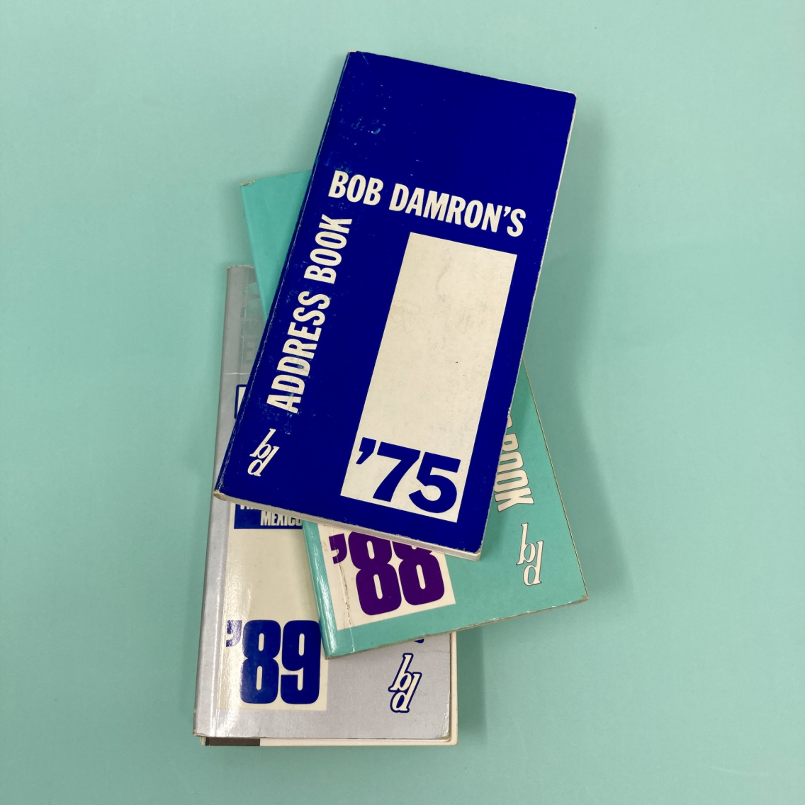 1975, 1988, and 1989 "Bob Damron's Address Book" stacked on a teal background