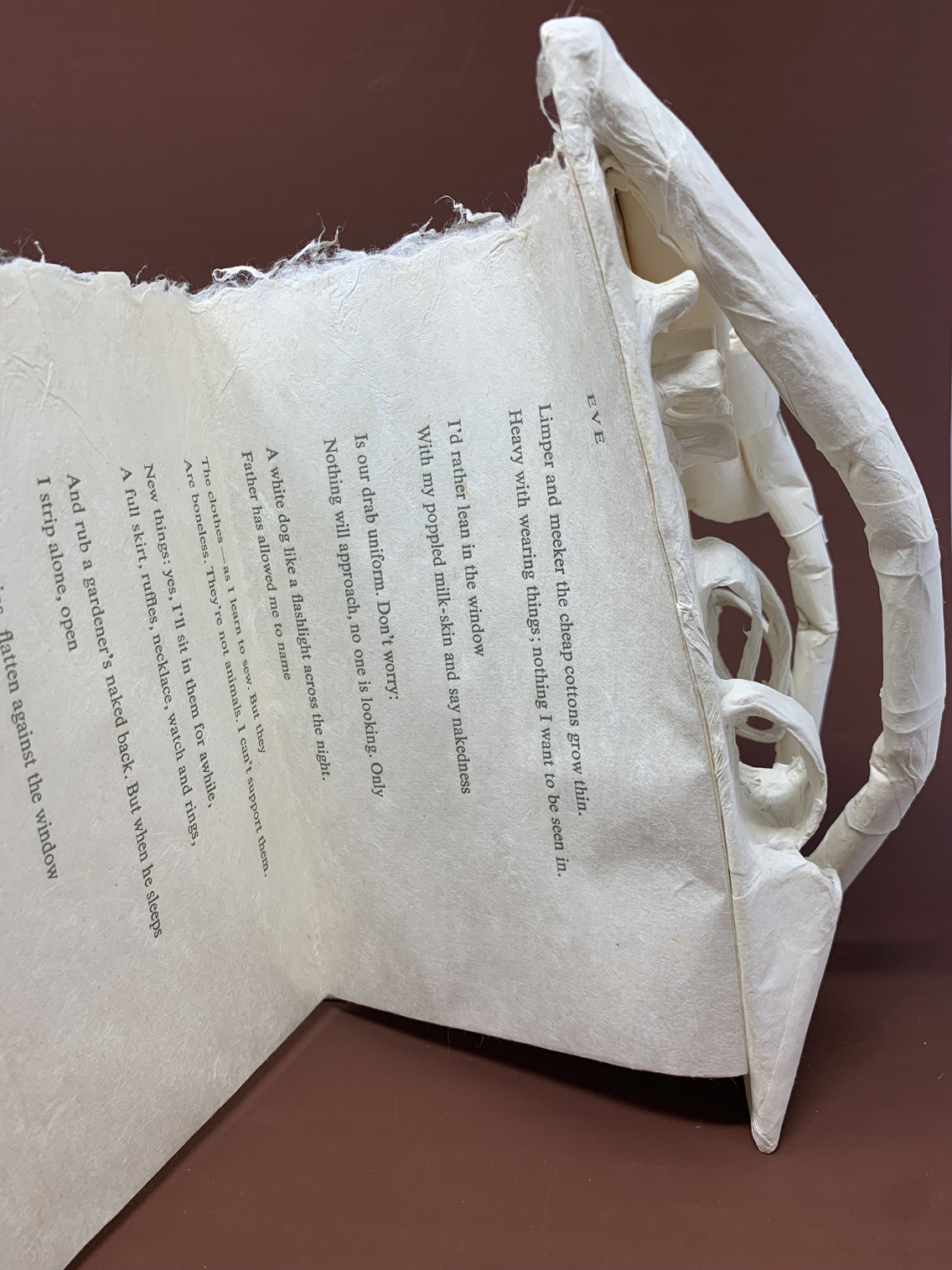 Close-up of a torn, folded white paper sculpture with printed poem text on a dark reddish-brown background.