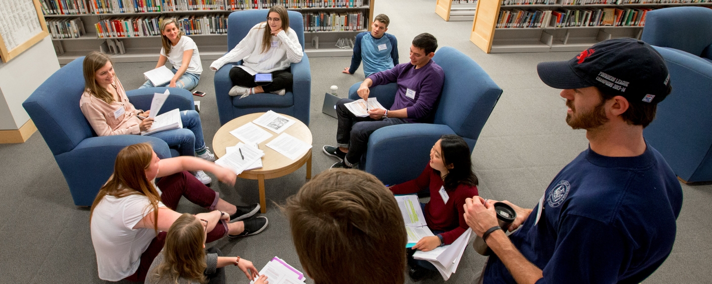 Group of ten male and female students sitting on chairs and on floor in library working.