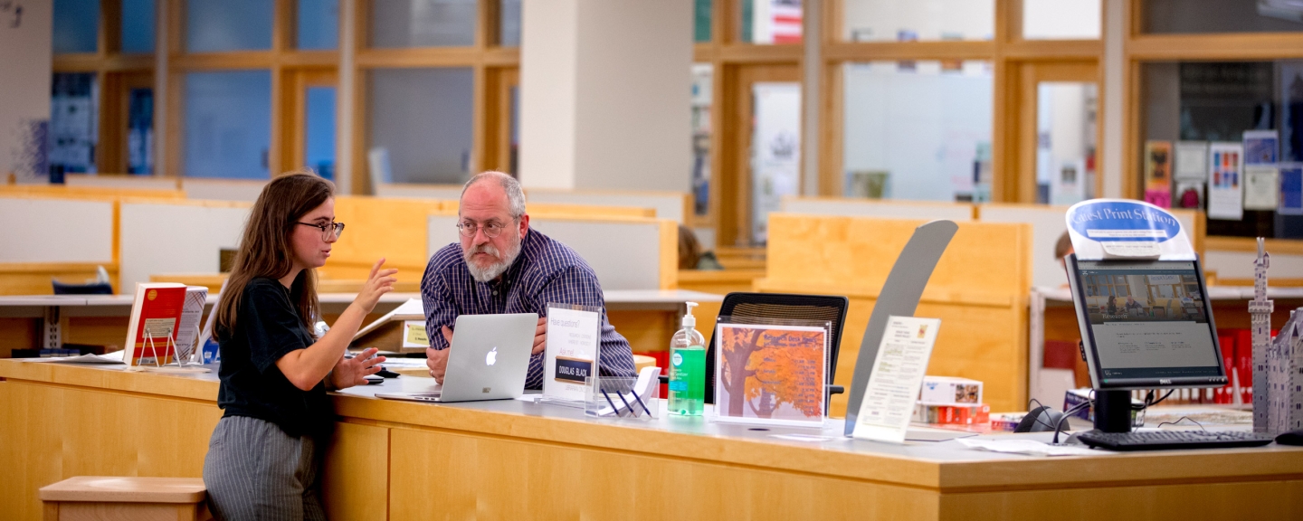 Female student getting guidance from male library employee at Research desk.