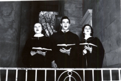three students dressed in black robes hold open books and stand on the steps of the chapel, mouths open in song