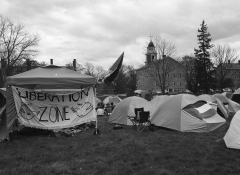 black and white photo of tents, camping chairs, flags, banner painted with a dove and text "liberation zone" on grassy lawn with Old Chapel in background