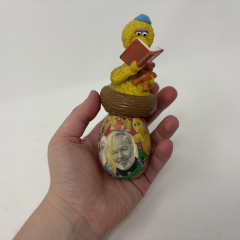 hand holds wooden egg decorated with a collage of photos of sesame street's big bird, a black and white photo of ron powers, and an oversized figurine of big bird sitting in a nest reading a book