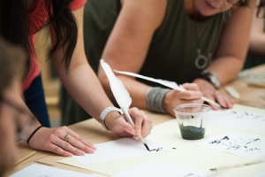 Two women hold feather quills and test writing with them during a workshop.