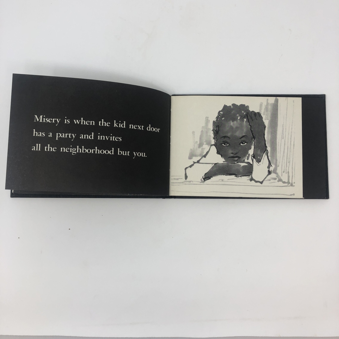 Photograph of a page in the children's book, Black Misery, by Langston Hughes and illustrated by Arouni. The text reads, "Misery is when the kid next door has a party and invites all the neighborhood but you." The accompanying illustration is of a Black child with their head in their hands, looking sadly up at the reader.