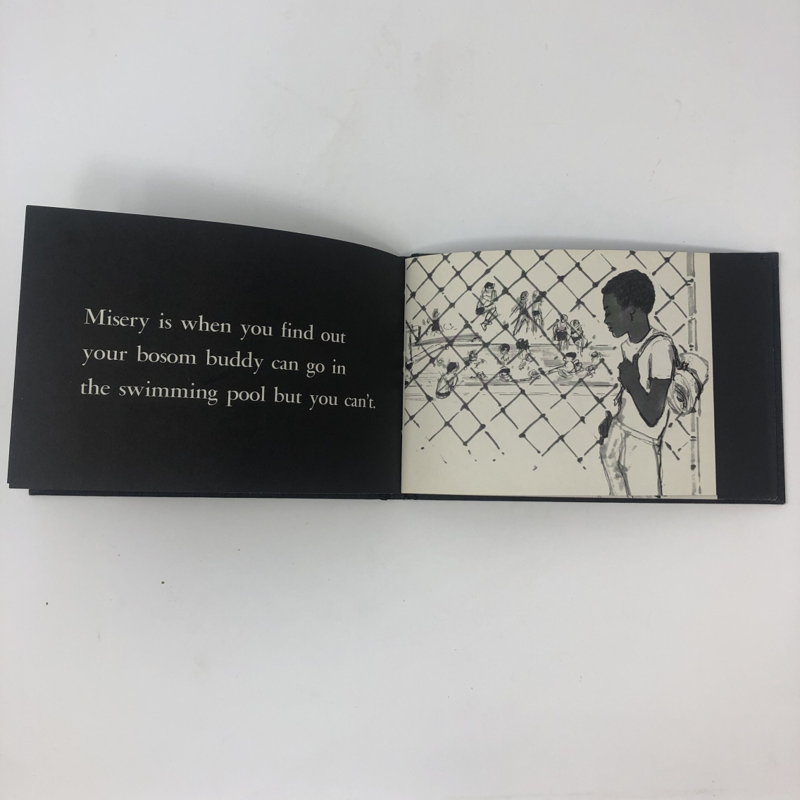Photograph of a page in the children's book Black Misery by Langston Hughes. The text reads, "Misery is when you find out your bosom buddy can go in the swimming pool but you can't." The illustration is of a Black child looking through a fence at other kids swimming in a pool.