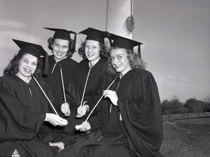 Black-and-white photograph of four female graduates wearing caps and gowns and holding long white pipes at commencement
