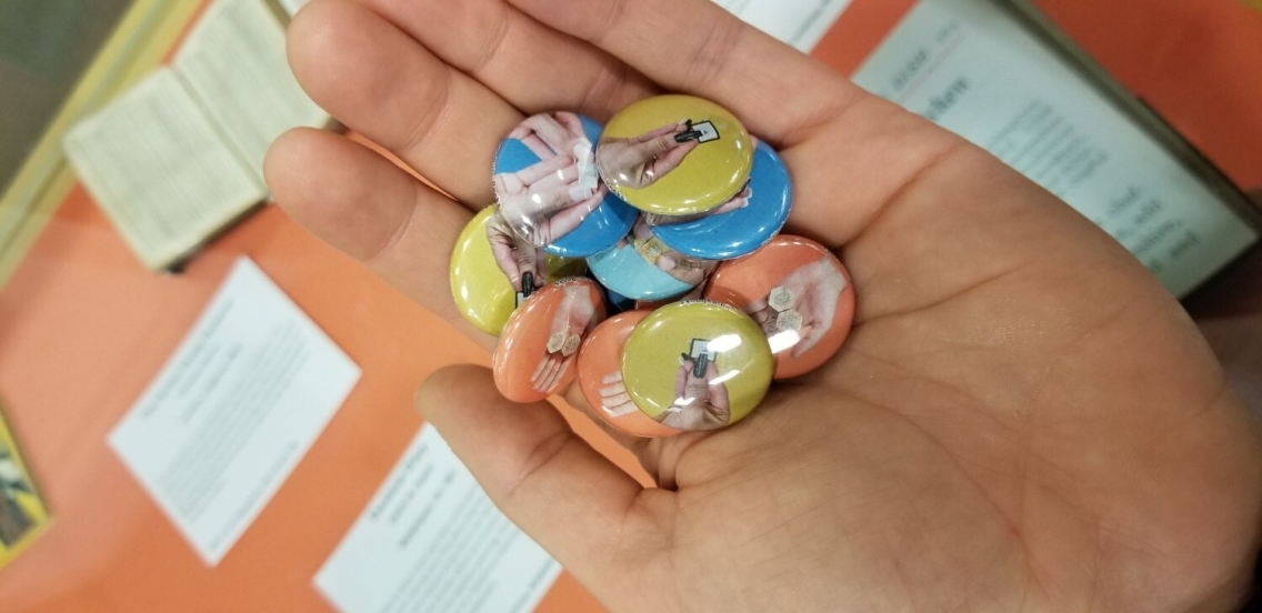 Hand holding pile of multi-colored pinback buttons featuring images of hands holding tiny books.