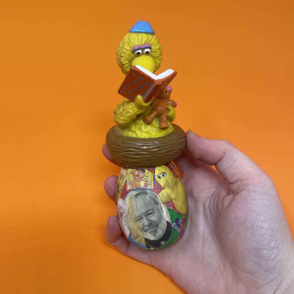 against an orange background, a white hand holding a painted and decorated wooden egg with Big Bird reading a book on top. 