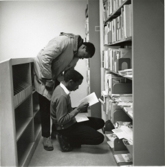 Black and white photograph of two students in the library looking at books on a shelf.