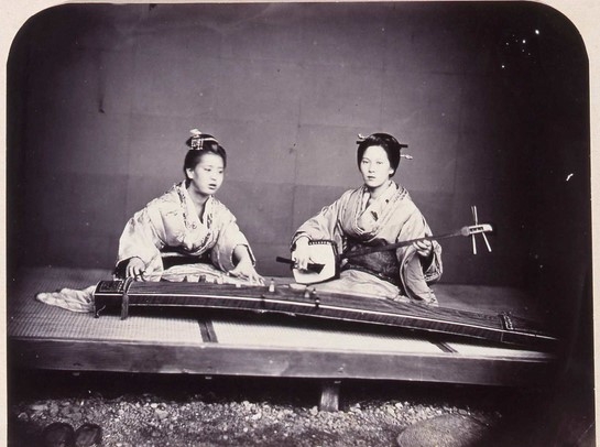 Archival photograph showing two Japanese women, one playing a Koto, and another playing Shamisen.