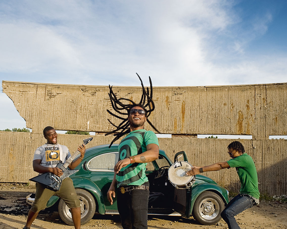 three members of a rock band play instruments and revel in front of a green Volkswagen Beetle
