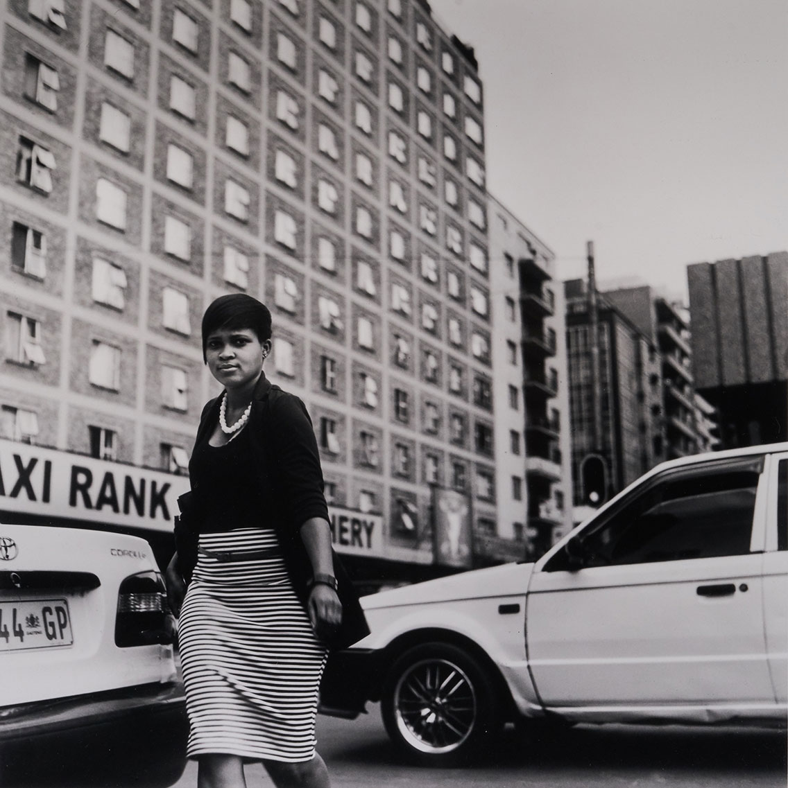 a woman wearing business attire walks between cars in front of alarge commercial building