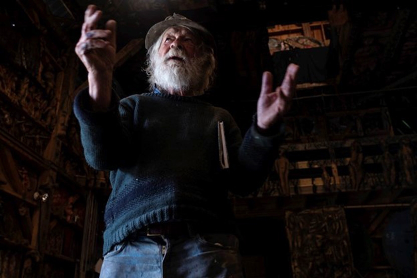 a gentleman with a white beard and clad in jeans and a blue sweater gestures toward people out of view behind the photographer