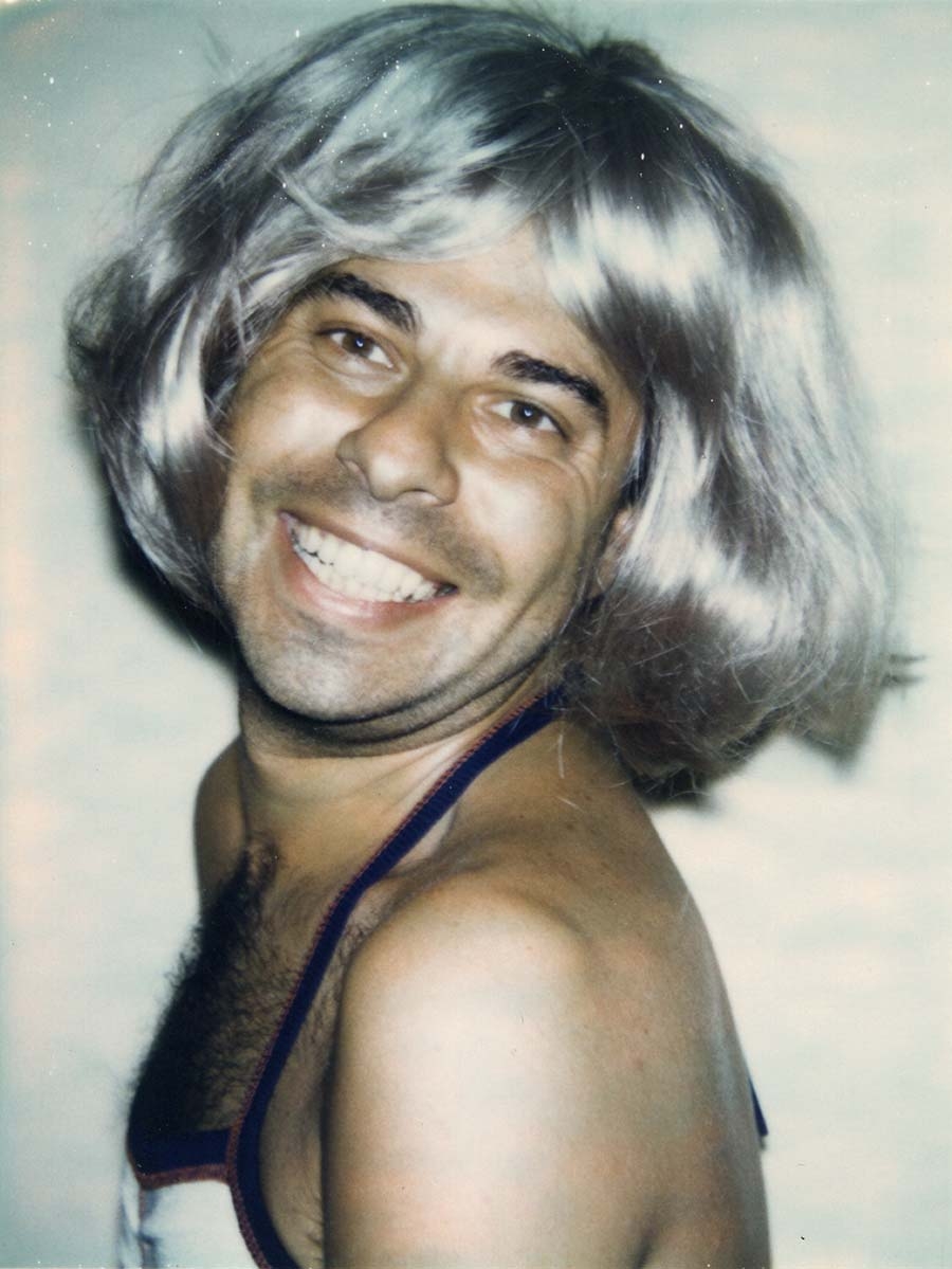 A smiling man with a hairy chest wears a silver bob-style wig