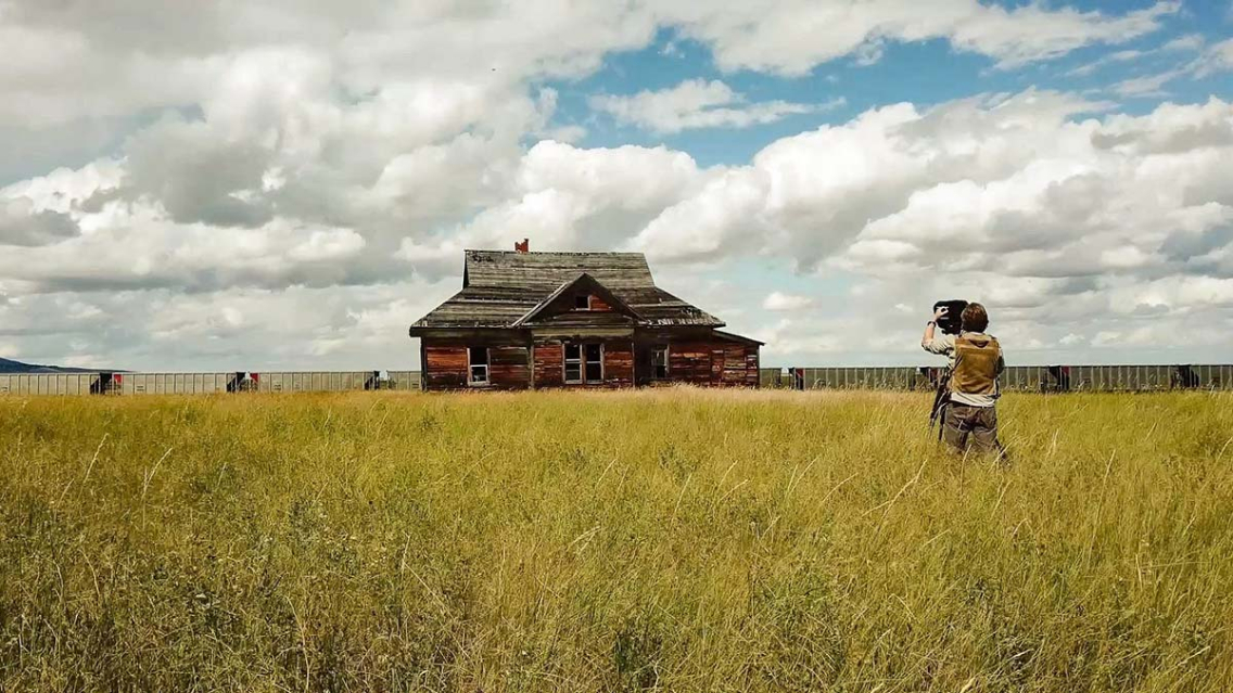 film still showing a filmmaker capturing footage of an abandoned farmhouse in a field of tall grass with a freight train passing in the background