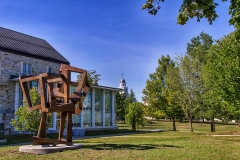 intersecting bronze cubes stacked and balanced, with a modern stone and glass building and blue sky in the background