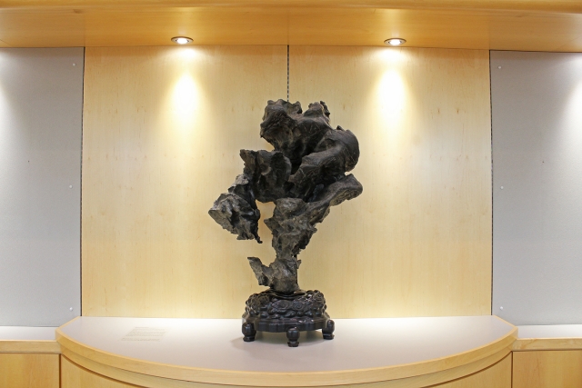 a dark gray carved piece of limestone with many nooks and crannies balances delicately atop a wooden base upon a counter with spotlights illuminating it from above
