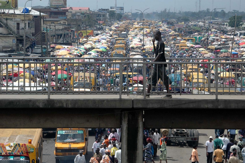 a black man talks on a cellphone as he walks across a bridge with a gridlock of traffic and pedestrians in the background