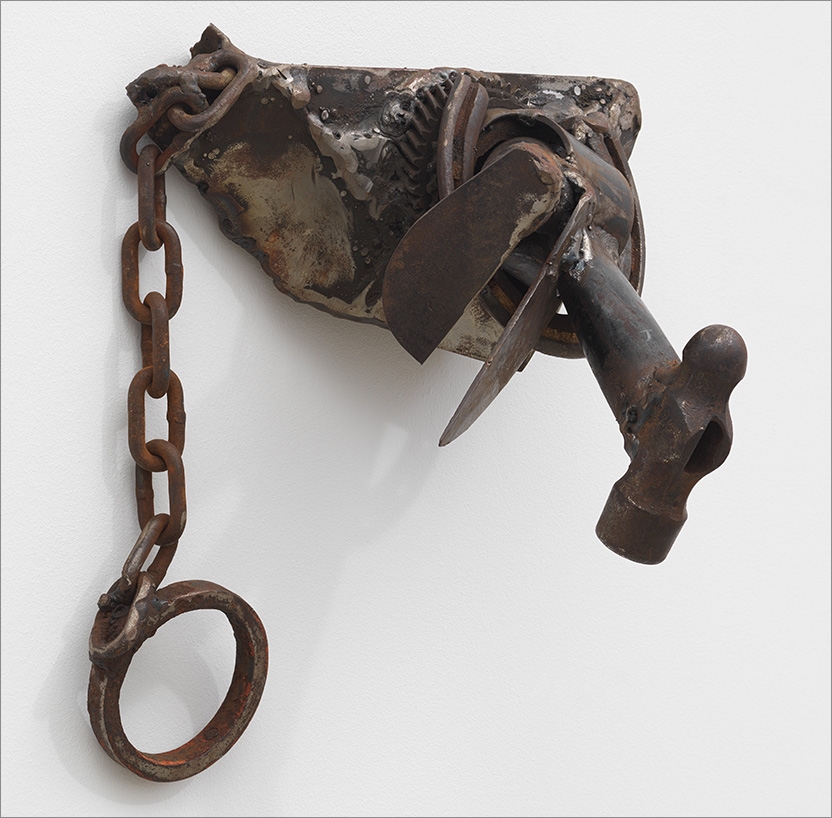 a collection of various metal elements - a hammer, chain links, gears, etc. - welded together in haphazard fashion and mounted to a white wall