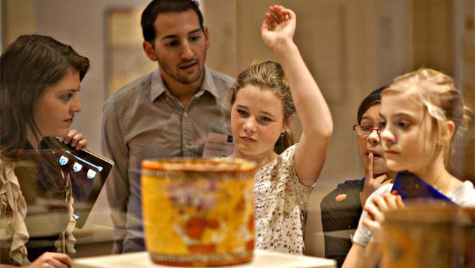 A young student raises her hand to ask about a ceramic vase they are viewing.