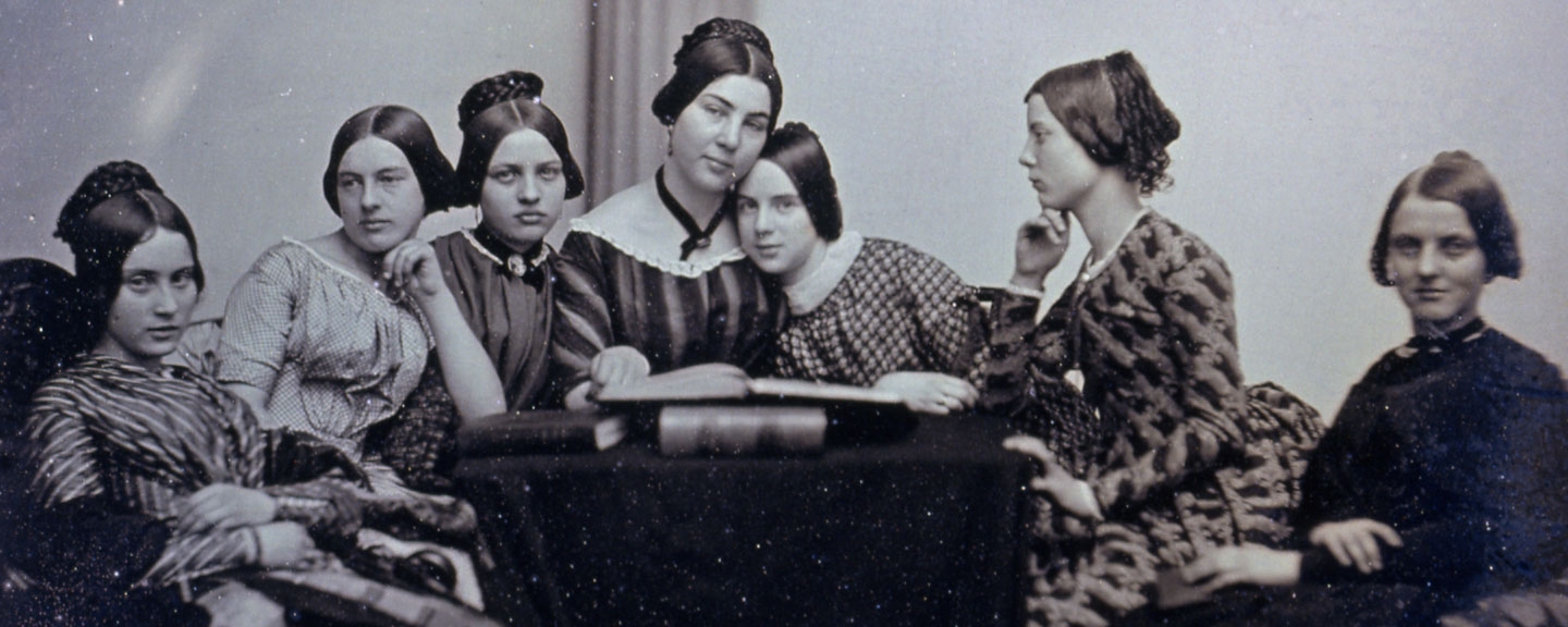 six female students sit around a table with their teacher, all wearing 19th century dress